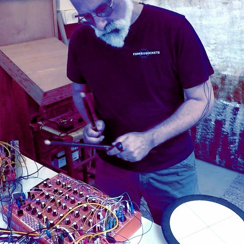 Desaturated photo of the artist bent over a desk, holding two drumsticks over a synth bearing a lot of knobs and wires. 