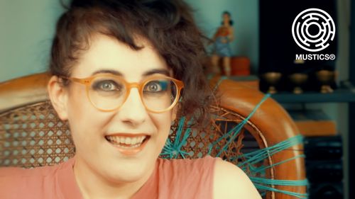 Meljoann sits on a wicker chair, repaired with green wool. A cheeky grin is evident. A statue of Wonder Woman is visible in the background.